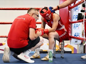 A boxer in red gear sits on a stool in a ring while a coach in a red shirt kneels beside them, and another person offers a towel.