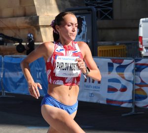 An athlete runs in a race, wearing a red and white top and blue shorts, with a bib displaying "PARLOV KOSTI" and "TARANTO 2026."